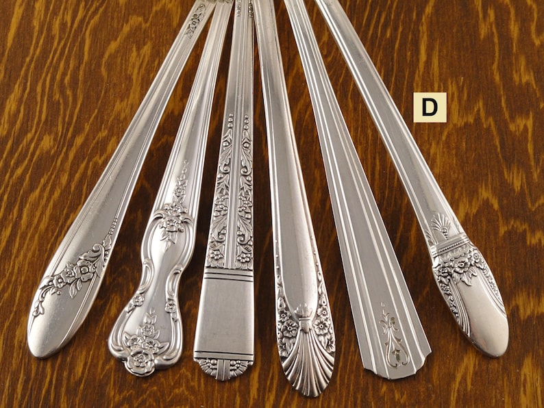 May include: Six silver-colored antique spoons arranged on a wooden surface. Each spoon features a unique decorative handle design, including floral and geometric patterns. The spoons are arranged in a fan shape, showcasing their varied styles.
