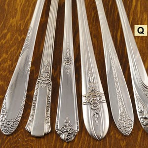 May include: Six antique silver spoons with ornate floral and scroll designs. Each spoon has a unique pattern, showcasing detailed craftsmanship. The spoons are arranged on a wooden surface, highlighting their polished finish and vintage appeal.