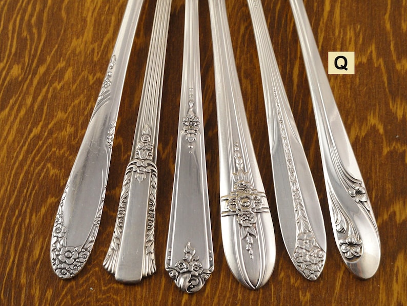 May include: Six antique silver spoons with ornate floral and scroll designs. Each spoon has a unique pattern, showcasing detailed craftsmanship. The spoons are arranged on a wooden surface, highlighting their polished finish and vintage appeal.