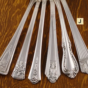 May include: An assortment of antique silver flatware, including forks and spoons, with ornate floral and geometric designs. The silverware is arranged on a wooden surface, showcasing the intricate details and polished finish of each piece.