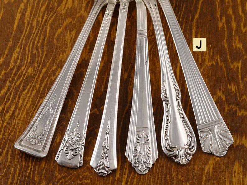 May include: An assortment of antique silver flatware, including forks and spoons, with ornate floral and geometric designs. The silverware is arranged on a wooden surface, showcasing the intricate details and polished finish of each piece.