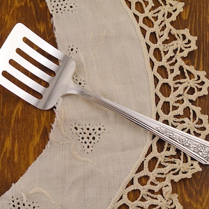 May include: A silver slotted spatula with a decorative handle, resting on a cream-colored lace doily. The spatula has four slots and a floral design on the handle. The background is a dark wood surface.