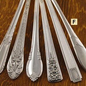 May include: An assortment of six silver spoons, each with a unique decorative handle design. The spoons are arranged on a wooden surface, showcasing various floral and geometric patterns. The spoons are made of a shiny, reflective metal.