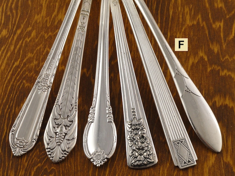 May include: An assortment of six silver spoons, each with a unique decorative handle design. The spoons are arranged on a wooden surface, showcasing various floral and geometric patterns. The spoons are made of a shiny, reflective metal.