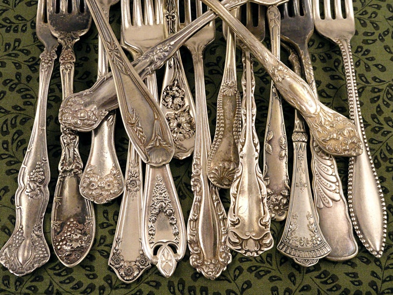 Puede incluir: Una colecci&oacute;n de tenedores de plata vintage con dise&ntilde;os ornamentados. Los tenedores est&aacute;n dispuestos en forma de abanico sobre una tela de estampado verde.