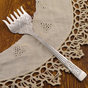 May include: A silver serving fork with a decorative floral design on the head and handle. The fork has six tines and is resting on a cream-colored lace doily. The background is a dark wooden surface.