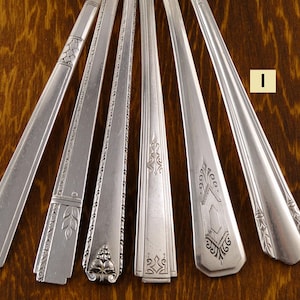 May include: A collection of vintage silver flatware, including spoons and forks, arranged on a wooden surface. Each piece features unique decorative patterns and designs, showcasing the craftsmanship of the era. The silverware is displayed in a fan-like arrangement.