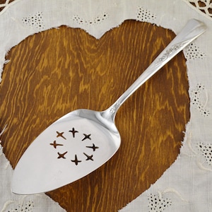 May include: A silver pie server with a decorative handle and a slotted blade featuring a pattern of crosses. The server rests on a wooden surface with a heart shape, and a white lace cloth.