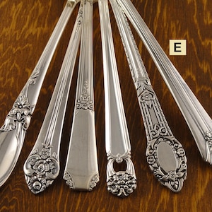 May include: A collection of six silver-colored spoons with ornate floral and scroll designs. Each spoon features a unique pattern on the handle, showcasing different styles of decorative silverware. The spoons are arranged on a wooden surface.