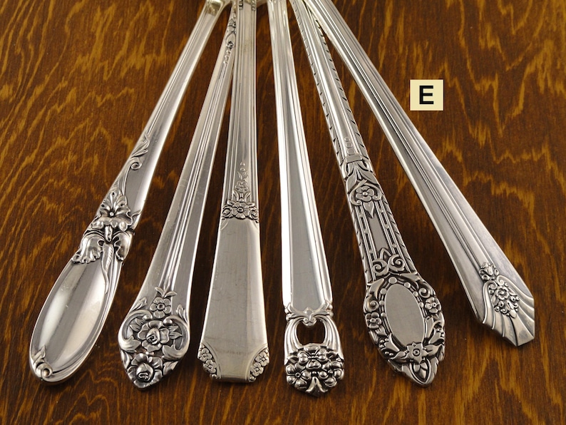May include: A collection of six silver-colored spoons with ornate floral and scroll designs. Each spoon features a unique pattern on the handle, showcasing different styles of decorative silverware. The spoons are arranged on a wooden surface.