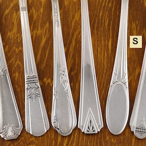 May include: Six silver spoons with different decorative patterns. The spoons are arranged side by side, showcasing various designs on their handles. The spoons are made of a shiny, reflective metal, and the background is a wooden surface.