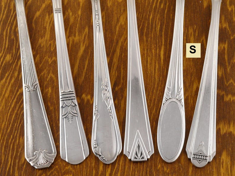 May include: Six silver spoons with different decorative patterns. The spoons are arranged side by side, showcasing various designs on their handles. The spoons are made of a shiny, reflective metal, and the background is a wooden surface.