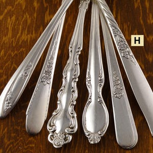 May include: Close-up of several silver spoons and serving utensils with ornate floral and scroll designs. The utensils are arranged on a dark wood surface, showcasing their intricate patterns and polished finish. The letter "H" is visible in the image.
