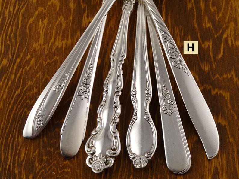 May include: Close-up of several silver spoons and serving utensils with ornate floral and scroll designs. The utensils are arranged on a dark wood surface, showcasing their intricate patterns and polished finish. The letter "H" is visible in the image.