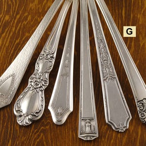 May include: An overhead shot showcases seven antique silver spoons, each with unique handle designs. The spoons are arranged on a wooden surface, with the letter "G" in a yellow square visible. The spoon designs include floral, geometric, and textured patterns.