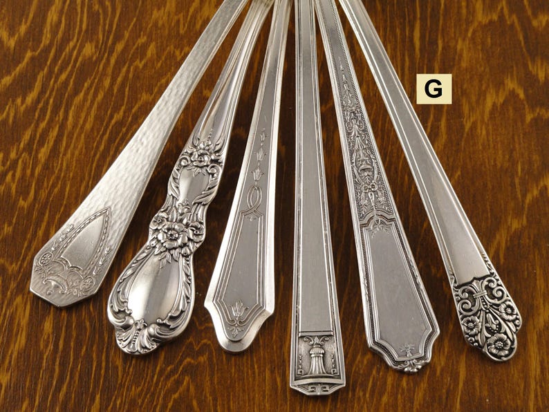 May include: An overhead shot showcases seven antique silver spoons, each with unique handle designs. The spoons are arranged on a wooden surface, with the letter "G" in a yellow square visible. The spoon designs include floral, geometric, and textured patterns.
