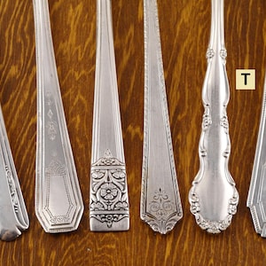 May include: Six silver spoons with ornate designs are displayed on a wooden surface. The spoons have different handle and bowl shapes, with floral and geometric patterns. The spoons are arranged in a row, showcasing their unique details and craftsmanship.