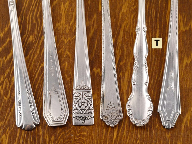 May include: Six silver spoons with ornate designs are displayed on a wooden surface. The spoons have different handle and bowl shapes, with floral and geometric patterns. The spoons are arranged in a row, showcasing their unique details and craftsmanship.