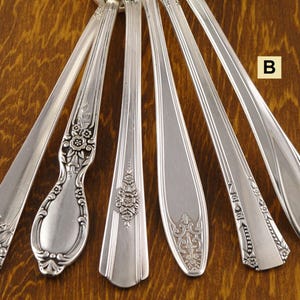 May include: Six antique silver spoons with various floral and ornate handle designs are arranged on a wooden surface. The spoons showcase different patterns, including detailed flower motifs and elegant curves, reflecting vintage craftsmanship.