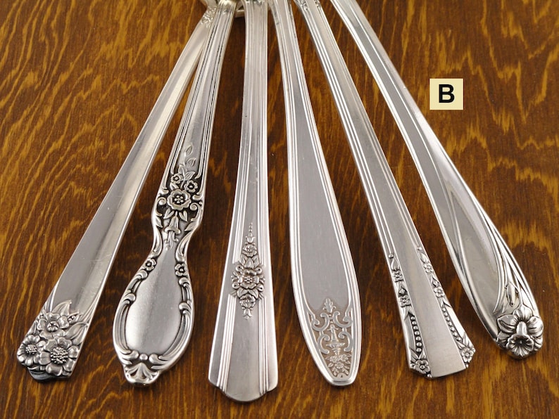 May include: Six antique silver spoons with various floral and ornate handle designs are arranged on a wooden surface. The spoons showcase different patterns, including detailed flower motifs and elegant curves, reflecting vintage craftsmanship.