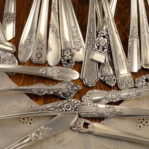 May include: A collection of vintage silver flatware with ornate handles. The silverware is arranged on a white lace tablecloth.