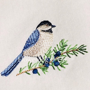 Puede incluir: Imagen bordada de un pájaro con plumas azules y beige posado en una rama verde con bayas azules. El pájaro tiene la cabeza negra y está sobre un fondo blanco. El texto "Flowerdog" es visible.