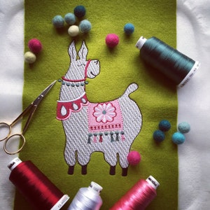 Op de afbeelding: Geborduurde lama met een roze zadel en wit en grijs bont op een groene vilt achtergrond. De lama is omringd door kleurrijke borduurgaren spoelen en kleine viltballetjes.