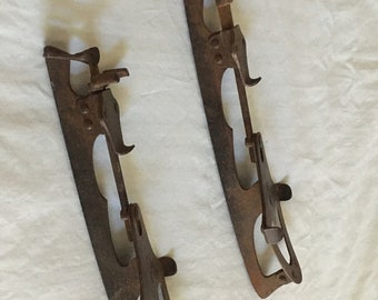 May include: Two antique ice skates made of rusty brown metal. Each skate has a blade, a heel clip, and a toe clip. The blades are long and narrow, with a curved shape. The skates are designed to be attached to boots for ice skating.