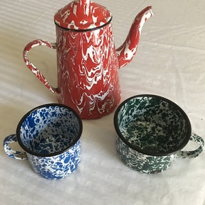 Könnte beinhalten: Eine rot-weiß emaillierte Kaffeekanne mit zwei passenden Tassen. Die Kaffeekanne hat einen schwarzen Rand und Griff. Eine Tasse ist blau-weiß, die andere grün-weiß. Das Set ist mit einem Marmormuster gestaltet.