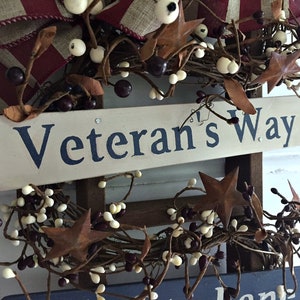 May include: A wooden sign with the words "Veteran's Way" painted in blue on a white background. The sign is decorated with a wreath of brown twigs, white berries, and rusty stars.
