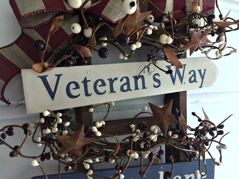 May include: A wooden sign with the words "Veteran's Way" painted in blue on a white background. The sign is decorated with a wreath of brown twigs, white berries, and rusty stars.