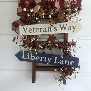 May include: A wooden ladder with a red, white, and blue ribbon and a garland of brown twigs and white berries. Two signs are attached to the ladder, one that reads "Veteran's Way" and the other that reads "Liberty Lane".