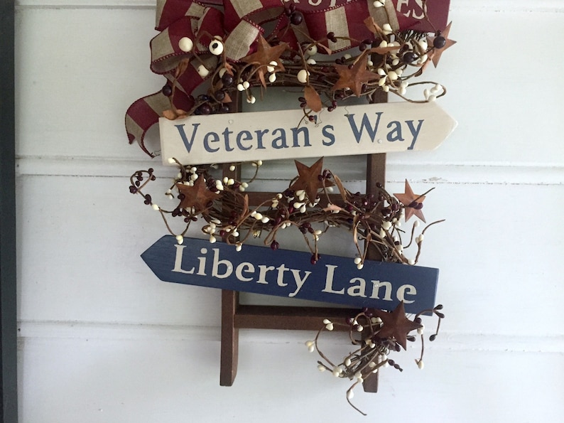 May include: A wooden ladder with a red, white, and blue ribbon and a garland of brown twigs and white berries. Two signs are attached to the ladder, one that reads "Veteran's Way" and the other that reads "Liberty Lane".