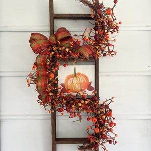May include: A rustic wooden ladder decorated with a fall-themed wreath. The wreath features orange and red berries, a plaid bow, and a sign with a pumpkin and the words "Hello Fall".