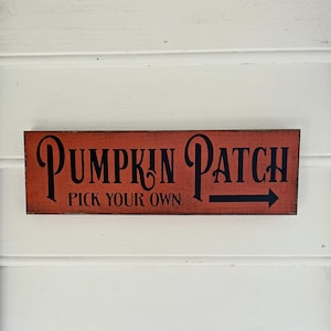 May include: A rectangular wooden sign with the words "PUMPKIN PATCH" in black script, with "PICK YOUR OWN" below. A black arrow points right. The sign has a distressed, orange-red background and is mounted on a white, horizontal-paneled wall.