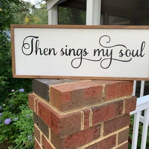 May include: A white wooden sign with a brown wood frame. The sign has black cursive lettering that reads "Then sings my soul".