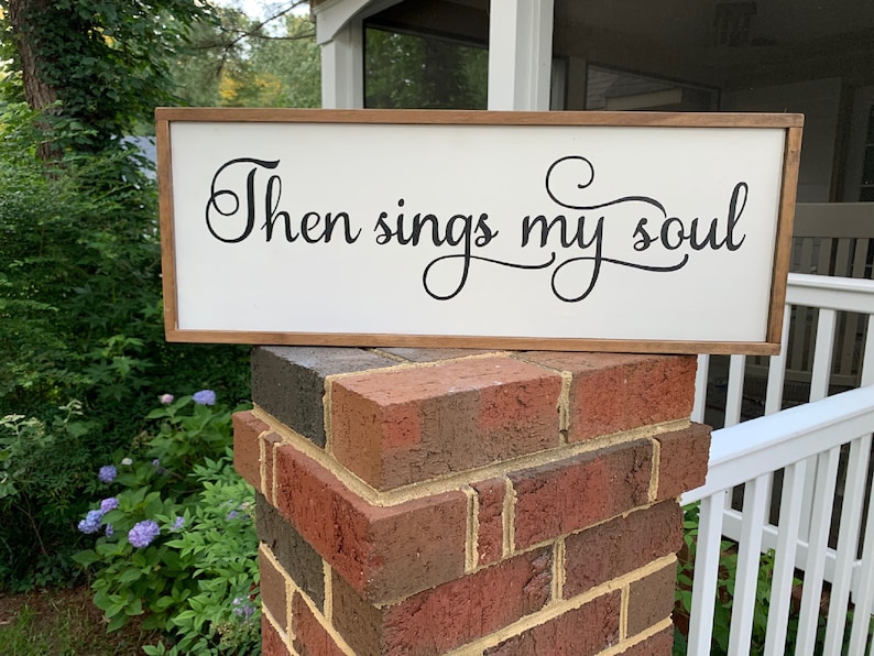May include: A white wooden sign with a brown wood frame. The sign has black cursive lettering that reads "Then sings my soul".