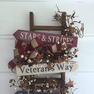 May include: A wooden ladder with two signs attached. The top sign is red and white and says "Stars & Stripes". The bottom sign is white and says "Veterans Way". The ladder is decorated with a red, white, and brown ribbon and white berries.