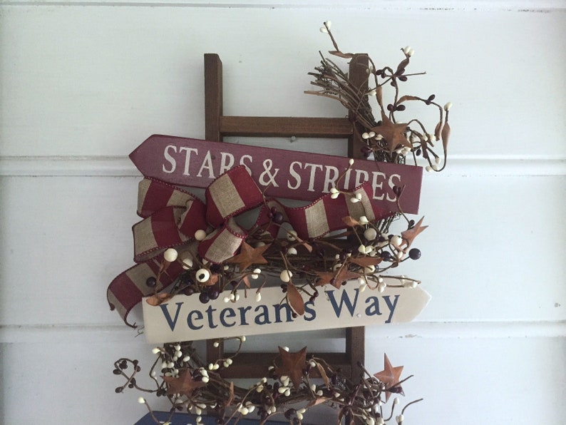 May include: A wooden ladder with two signs attached. The top sign is red and white and says "Stars & Stripes". The bottom sign is white and says "Veterans Way". The ladder is decorated with a red, white, and brown ribbon and white berries.