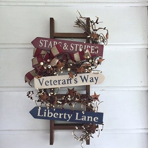 May include: A wooden ladder with three signs attached. The signs are painted in red, white, and blue with the text "Stars & Stripes", "Veteran's Way", and "Liberty Lane". The ladder is decorated with white berries and brown twigs.