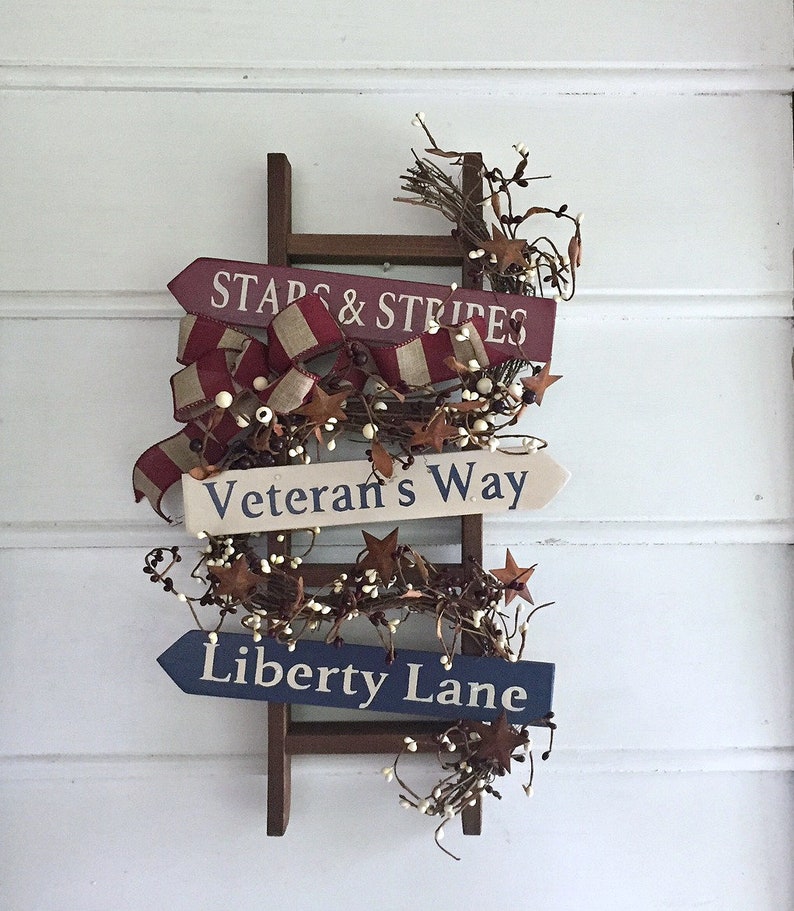 May include: A wooden ladder with three signs attached. The signs are painted in red, white, and blue with the text "Stars & Stripes", "Veteran's Way", and "Liberty Lane". The ladder is decorated with white berries and brown twigs.