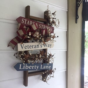 May include: A wooden ladder with three signs attached. The top sign reads "Stars & Stripes", the middle sign reads "Veteran's Way", and the bottom sign reads "Liberty Lane". The ladder is decorated with white berries and red, white, and blue ribbon.
