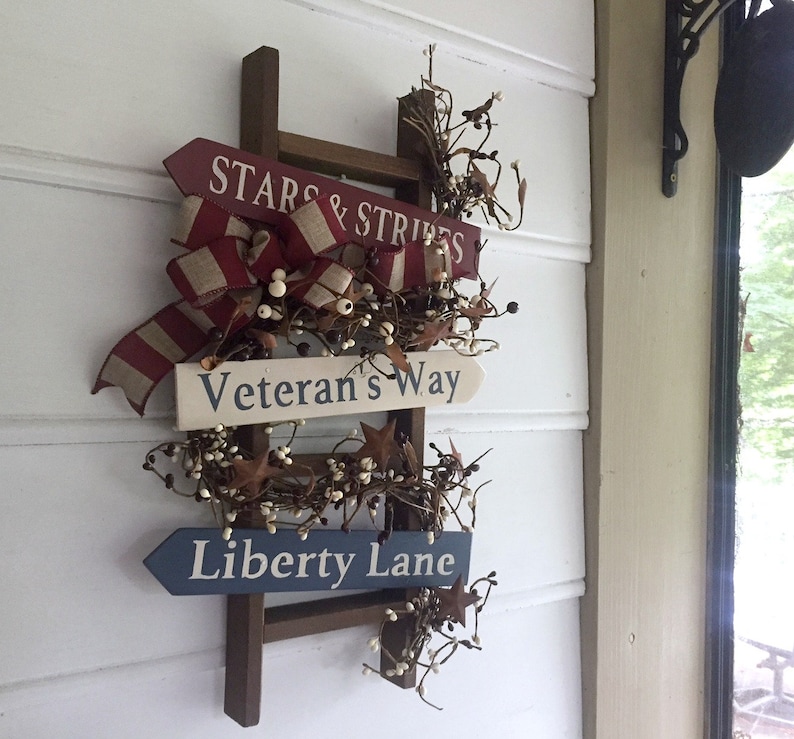 May include: A wooden ladder with three signs attached. The top sign reads "Stars & Stripes", the middle sign reads "Veteran's Way", and the bottom sign reads "Liberty Lane". The ladder is decorated with white berries and red, white, and blue ribbon.