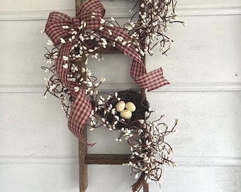 May include: A rustic wooden ladder decorated with a garland of brown twigs and small white berries. A red and white gingham ribbon is tied in a bow on the left side. A small dark brown bird nest containing three speckled cream eggs is attached.