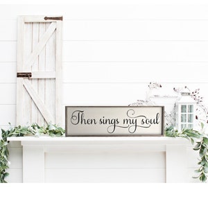 May include: A wooden sign with the text "Then sings my soul" in black cursive lettering. The sign is on a white mantel with a white door and greenery on either side.