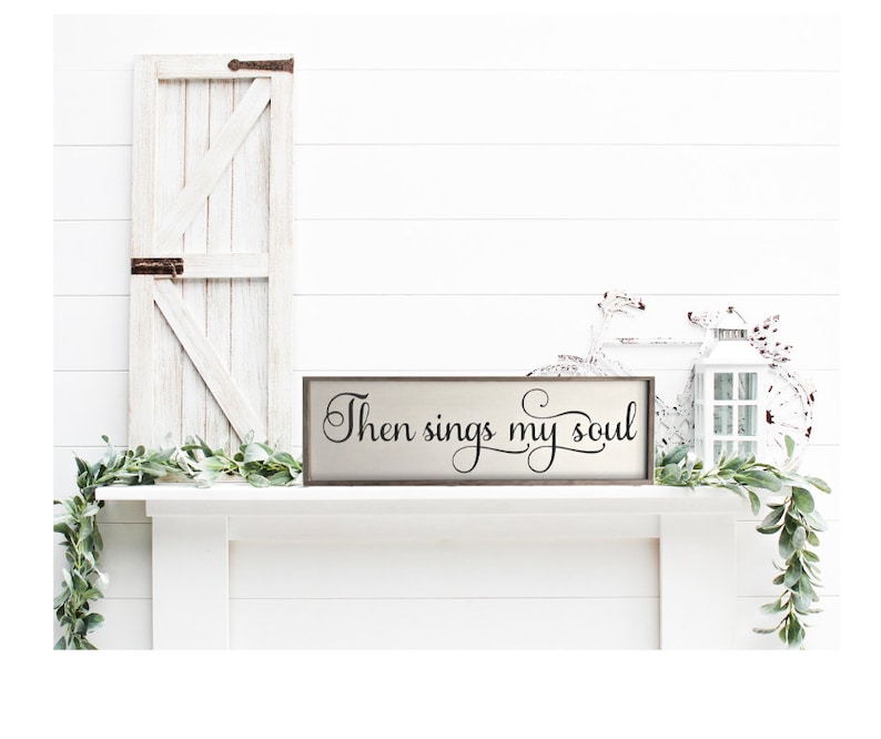 May include: A wooden sign with the text "Then sings my soul" in black cursive lettering. The sign is on a white mantel with a white door and greenery on either side.