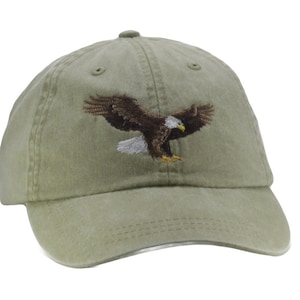 May include: Olive green baseball cap with an embroidered eagle design. The eagle is depicted with outstretched wings, featuring brown, white, and yellow details. The cap has a curved brim and a fabric strap closure.