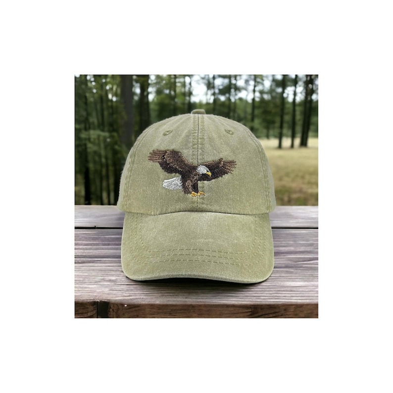 May include: Olive green baseball cap with an embroidered eagle in flight. The eagle has brown and white feathers and a yellow beak. The cap is made of a soft, worn fabric and has a curved brim. A casual accessory.