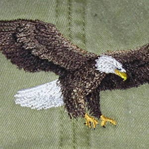 May include: Embroidered image of a bald eagle in flight, with outstretched wings. The eagle features brown and white feathers, a yellow beak and talons, and is set against a textured olive-green background. The embroidery detail is intricate.