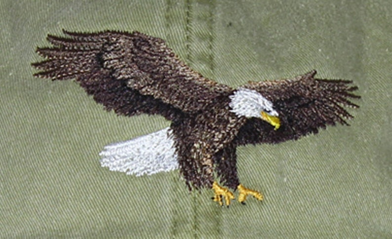 May include: Embroidered image of a bald eagle in flight, with outstretched wings. The eagle features brown and white feathers, a yellow beak and talons, and is set against a textured olive-green background. The embroidery detail is intricate.